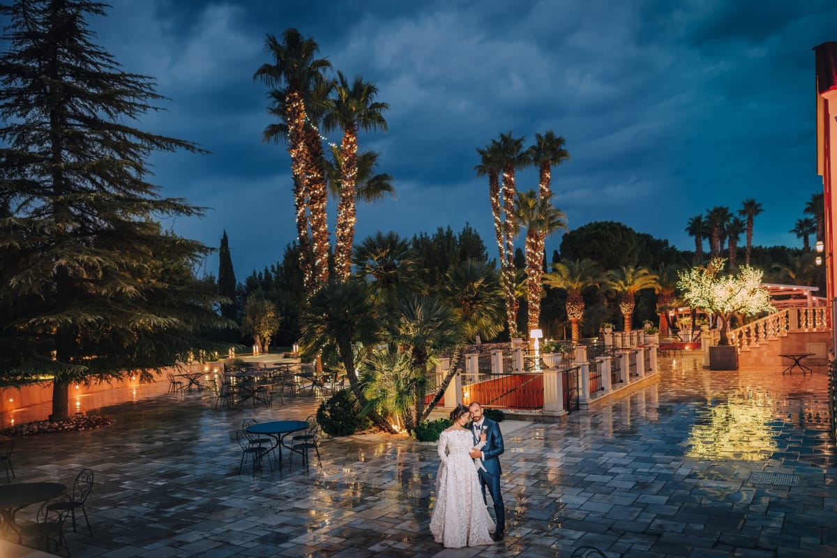 Foto serale di sposi all'aperto con pavimento bagnato dalla pioggia e palme illuminate.