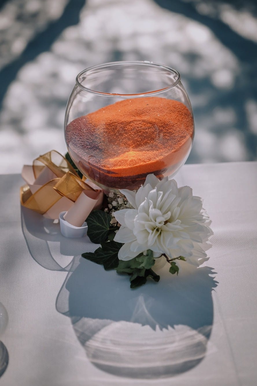Ampolla trasparente con sabbia arancione su tavola bianca con un fiore.