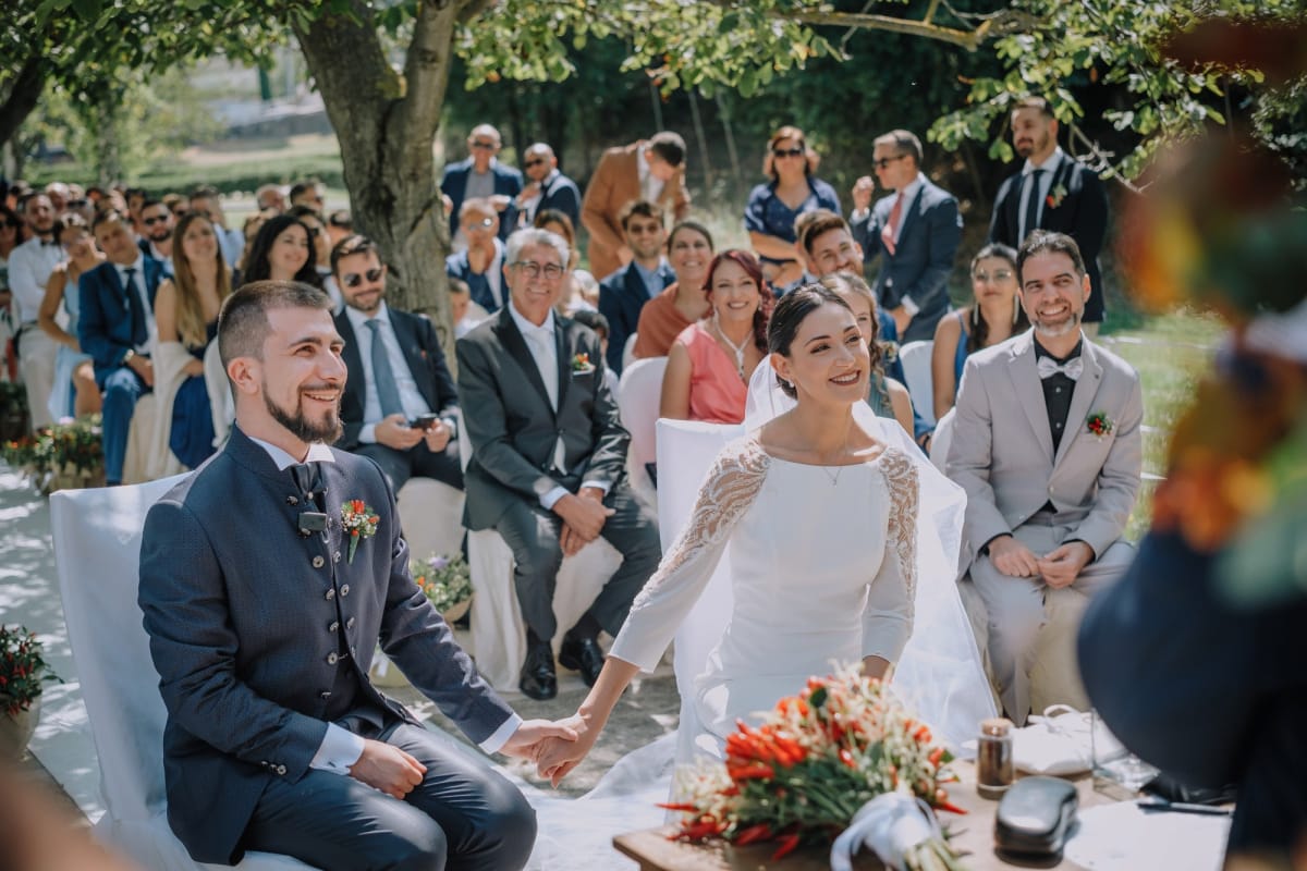 Coppia di sposi all'aperto si tiene per mano con invitati sullo sfondo.