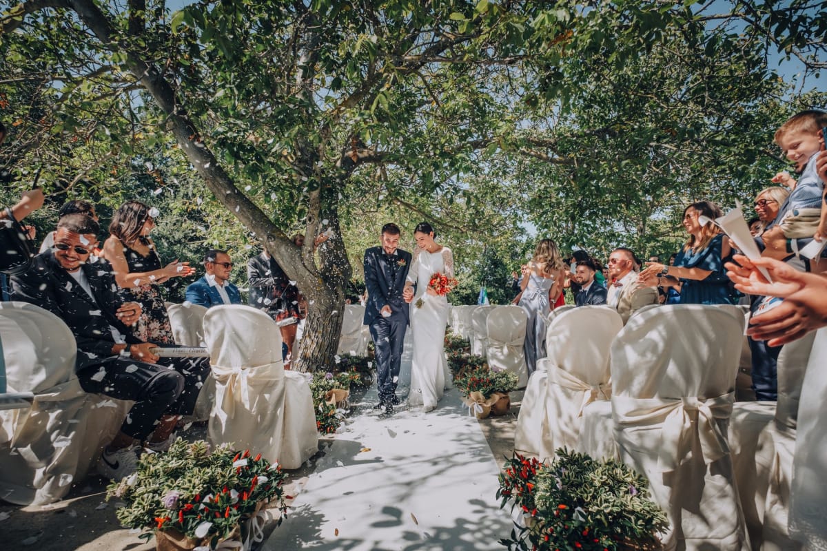 Coppia di sposi sfilano su tappeto bianco sotto un albero tra gli invitati.