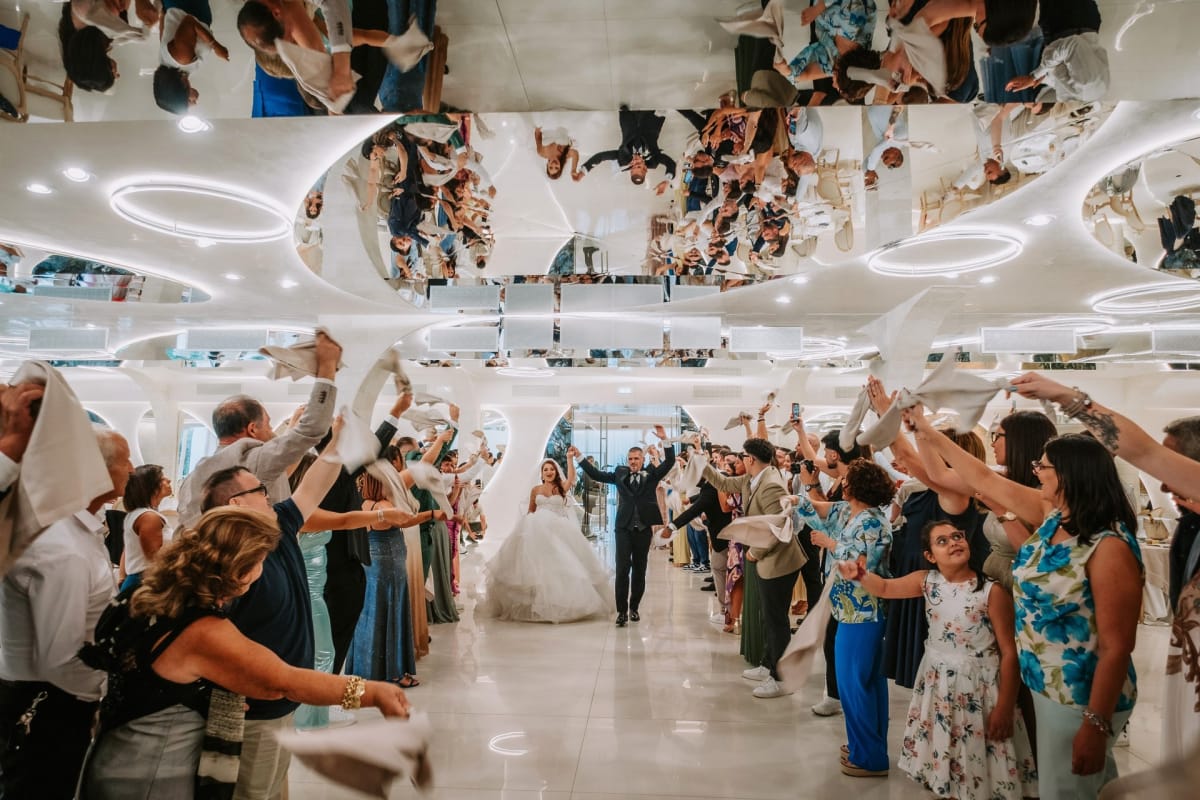 Gli sposi entrano in sala con le braccia alzate tra due ali di invitati che sventolano tovaglioli, riflessi nel soffitto a specchio