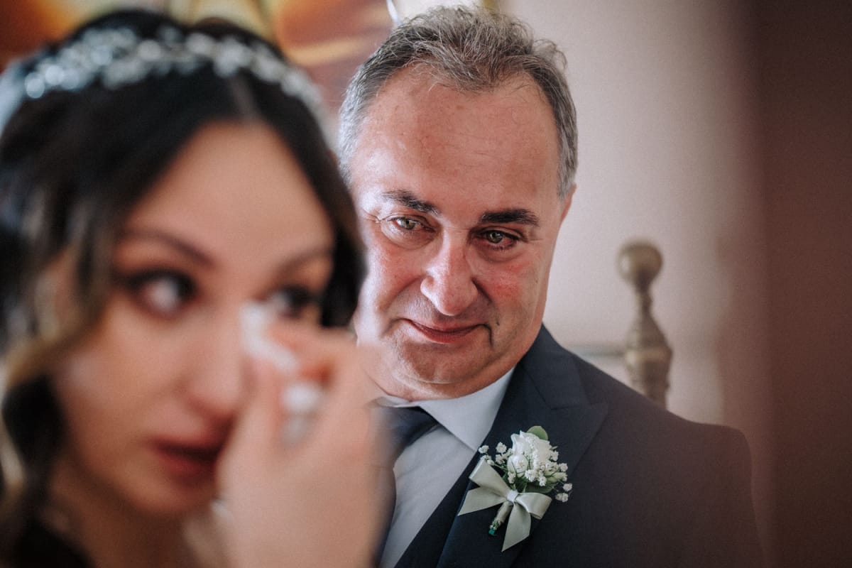 Il padre della sposa la guarda con gli occhi lucidi mentre lei, in primo piano, si asciuga una lacrima