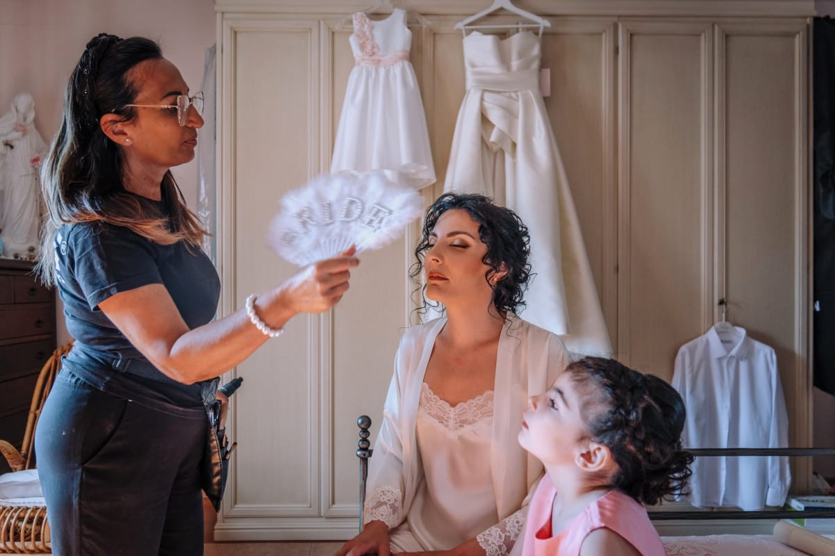 La sposa in vestaglia si fa truccare, rinfrescata con un ventaglio, mentre una bambina la osserva