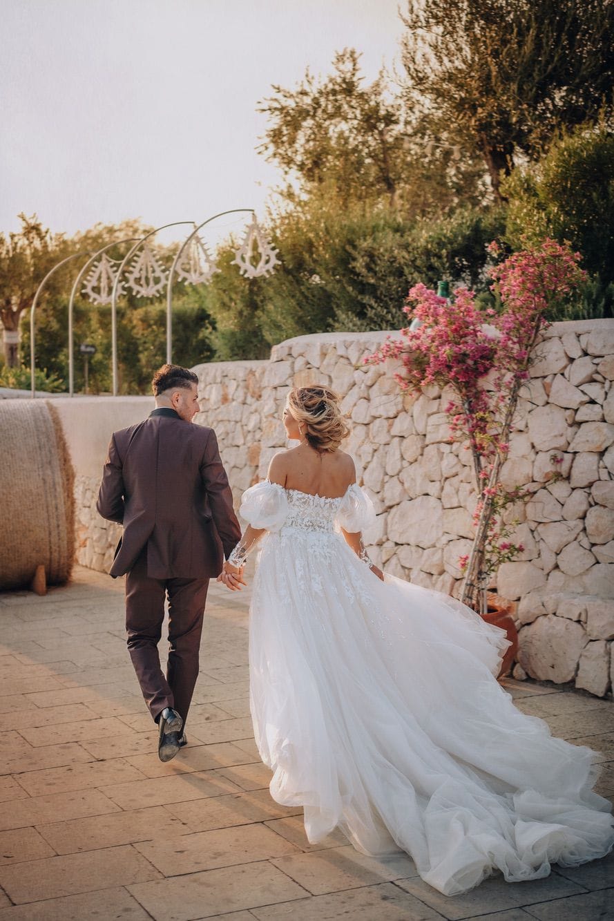 Gli sposi si allontanano mano nella mano lungo un vialetto in pietra, lei con lo strascico in tulle che si apre alle spalle, nella luce calda del tramonto