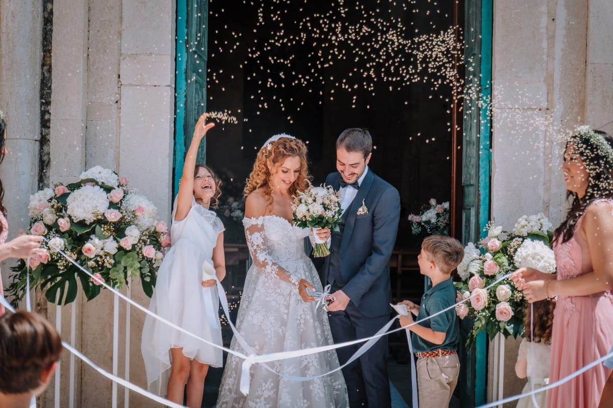Gli sposi escono dalla chiesa sotto il lancio del riso, tra bambini e invitati che li festeggiano.